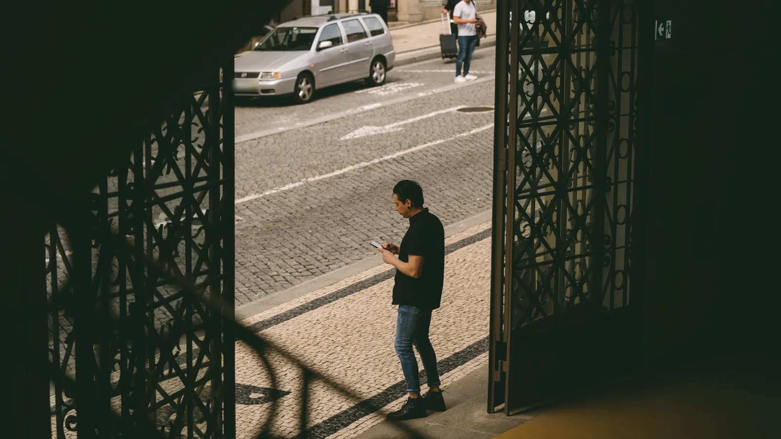 Homem a consultar o telemóvel num passeio de calçada portuguesa, à porta de um edifício com portões de ferro forjado, com um carro parado na rua ao fundo