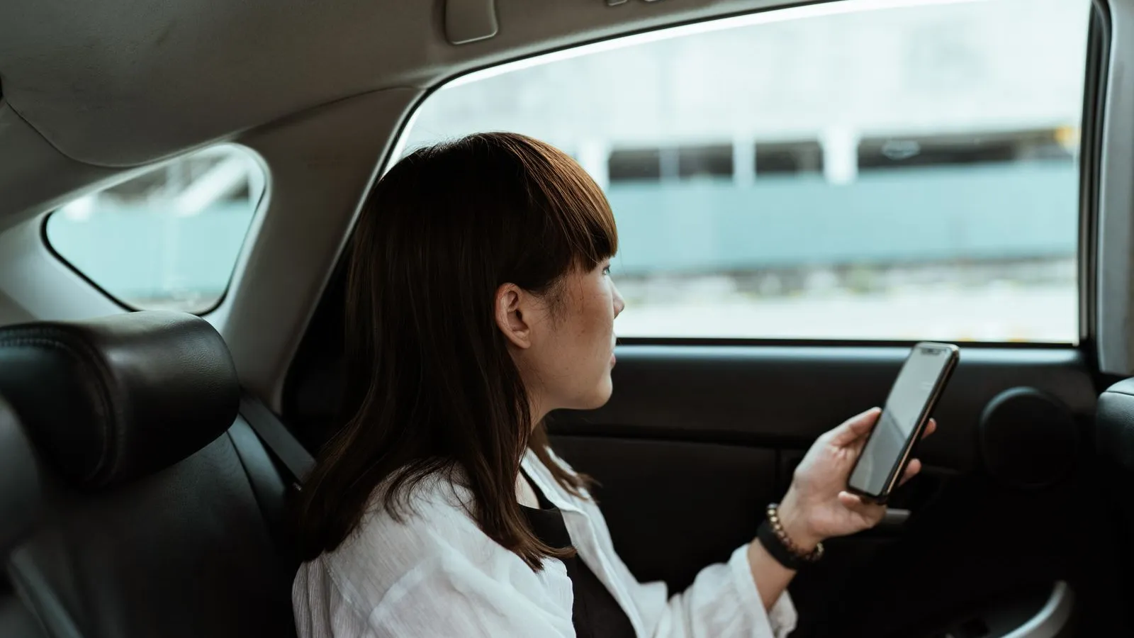 Mulher sentada no banco de trás de um carro, tranquila, com o telemóvel na mão a olhar pela janela.